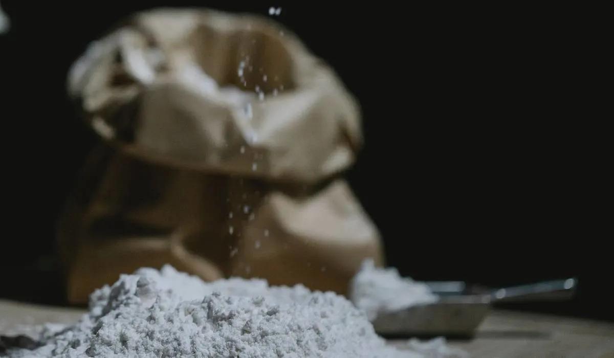 A pile of flour on a table with a cup and a bag in the backgroud.
