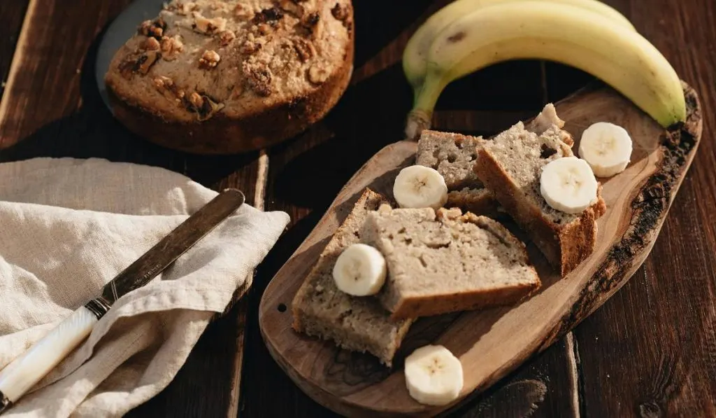 Banana bread on a wooden plate on table with a fresh banana on top of it.