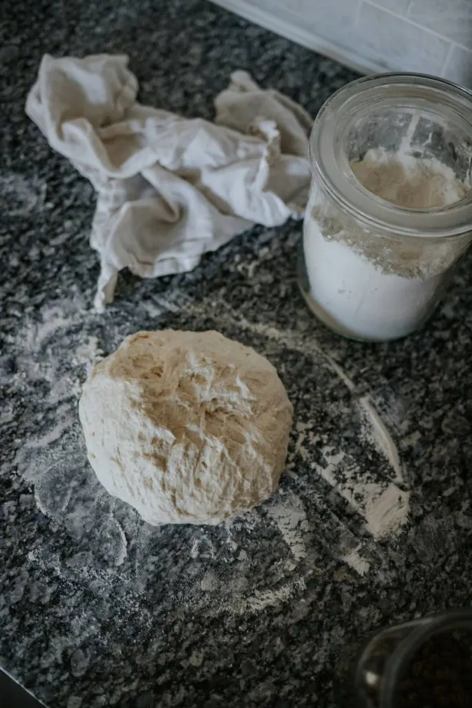 Dough on a table with a jar of flour and a cloth.
