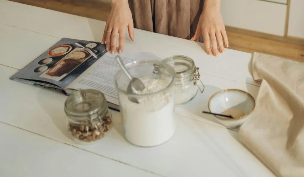 Woman reading bread cookbook with jars of ingrediens on a table. Best sourdough cookbooks